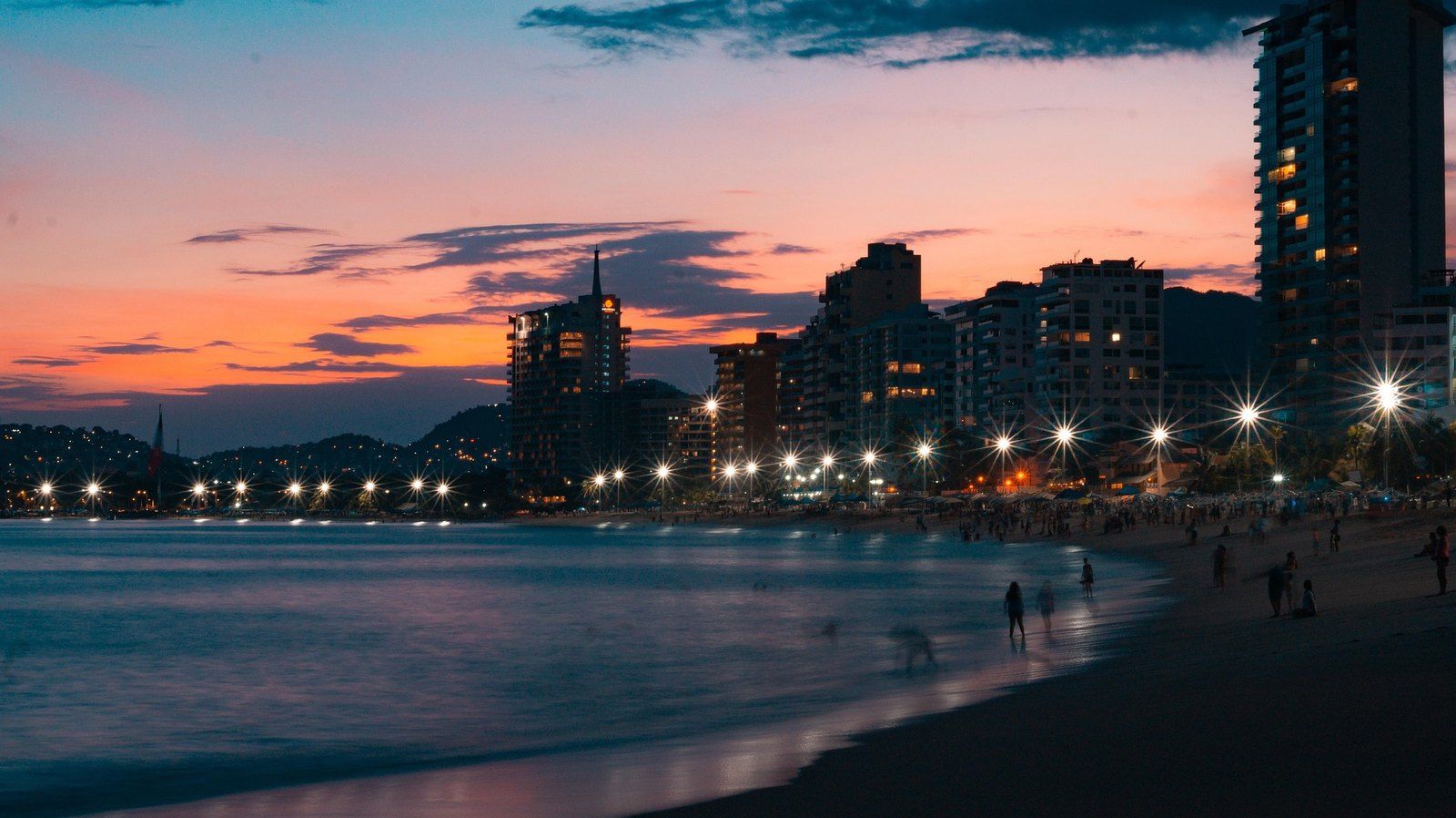 Acapulco Bay at sunset, with lit high-rise buildings along the beach under an orange sky
